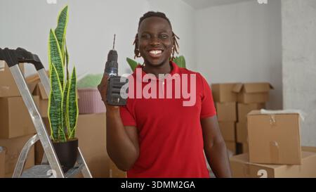Man holding drill beside ladder and potted plant in building cluttered with moving boxes and stacked cartons, smiling broadly; home improvement joy. Stock Photo