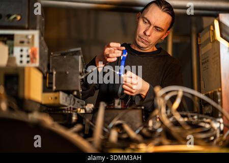 A male audio engineer repairs musical equipment in a workshop. A male worker repairs equipment by wrapping blue electrical tape around a wire in a wor Stock Photo