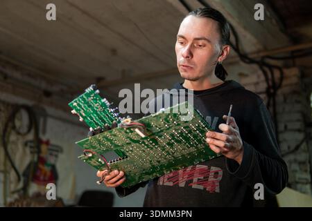 A male worker examines a circuit board for repair in a workshop setting. Stock Photo