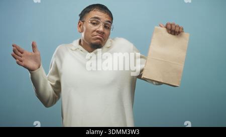Young hispanic man wearing glasses and cream sweater holding a crumpled paper bag while shrugging with hands in studio; uncertainty. Stock Photo