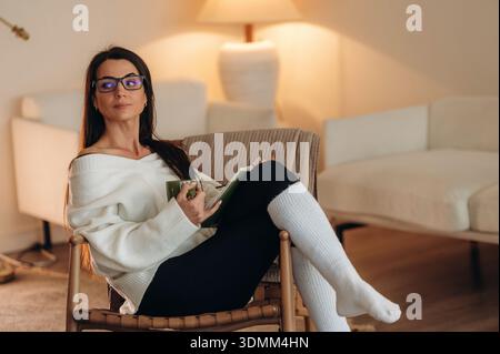 Smart person, in glasses. Relaxed woman is sitting in the chair and reading the book at home. Stock Photo