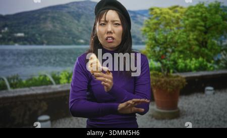Woman in purple turtleneck holds croissant with a grimace, pushes hand away and shrugs slightly on lakeside terrace; disgust rejection. Stock Photo