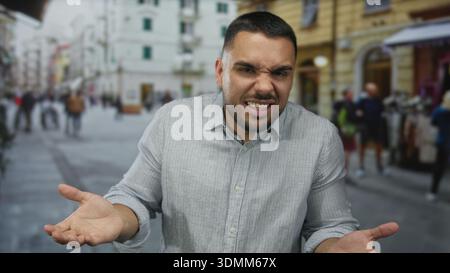 Hispanic young man showing open palms in striped grey shirt with confused expression on sunlit busy city street; confusion. Stock Photo