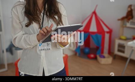 Hispanic woman using tablet in kindergarten classroom surrounded by colorful toys showcasing a preschool learning environment indoors. Stock Photo