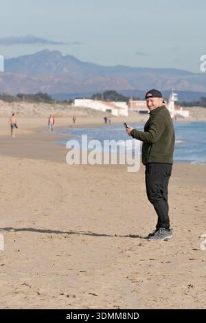 Man standing on a beach holding a smartphone with people walking and buildings near the water against a mountain horizon Stock Photo