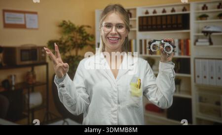 Woman optometrist wearing white coat and glasses holding trial frame and flashing peace sign while smiling in studio; friendly care. Stock Photo