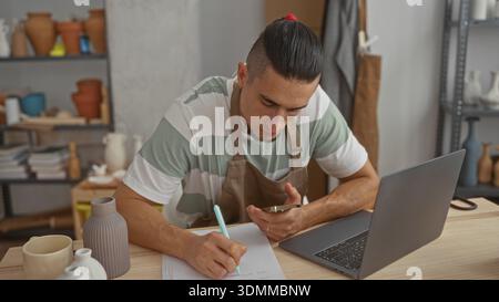 Man in apron holding phone and writing with hand on notepad beside laptop and ceramic vases on shelves in pottery studio; focused creativity. Stock Photo