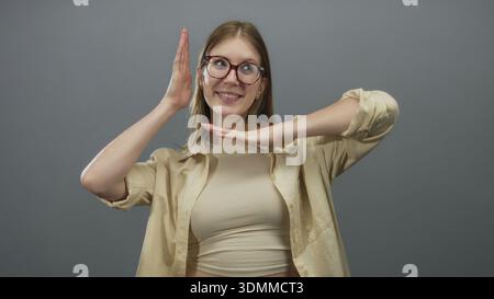 Young blonde woman wearing glasses smiles while framing face with hands in grey studio; playful joy. Stock Photo
