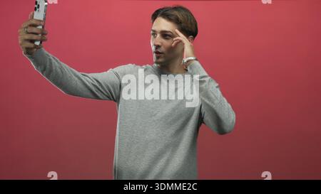 Man points finger at phone while holding smartphone at arm's length, wearing grey sweater in studio; confidence connection. Stock Photo