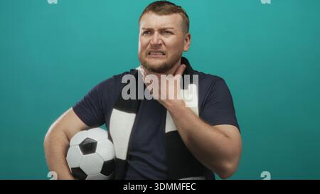 Man holding soccer ball while wearing black and white fan scarf, clutching throat with pained expression in teal studio backdrop; pain. Stock Photo