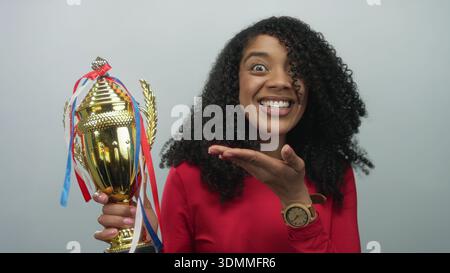 African american woman firmly holds golden trophy with ribbons and raises fist in light grey studio; joy victory. Stock Photo