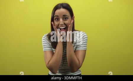 Woman touching cheeks and smiling wearing green striped tshirt in studio with yellow backdrop; surprise reaction moment. Stock Photo