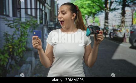 Woman holding game controller and creditcard, smiling with eyes closed and playful expression on city street between buildings; joy purchase. Stock Photo
