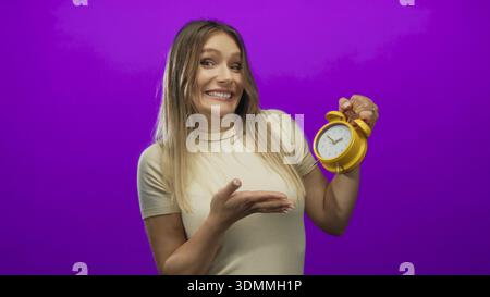 Woman holding yellow alarm clock against vibrant purple background with a confused expression, showcasing time management concept with youthful casual Stock Photo