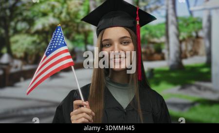 Woman holding flag in park, smiling in black graduation cap and gown with red tassel, standing on green lawn by benches; pride. Stock Photo