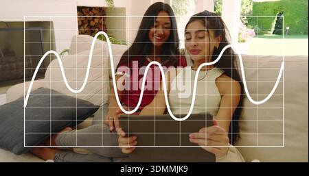 Watching two women sitting close on sofa at home, holding tablet with data overlay Stock Photo