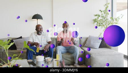 Smiling senior holding drink and son in coral tee reacting on gray sofa, with floating spheres Stock Photo