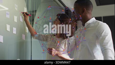 Writing colleagues in business attire collaborating at glass wall in office, Post-its and hologram Stock Photo