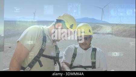 Two technicians examining tablet on arid plain near turbines, in yellow hard hats, harnesses, HUD Stock Photo