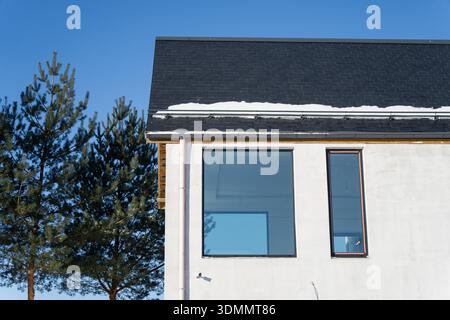 Minimalist home exterior with dark roof tiles, snow on eaves, modern glazing and vertical window, clear winter sky and pine tree silhouettes Stock Photo