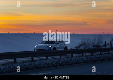 Winter road travel scene with white SUV and lit headlights, guardrail and wet asphalt, wide snow field under orange twilight sky with distant posts Stock Photo