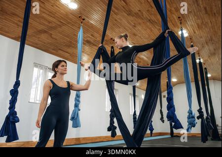 Teaching, support conception. Female instructor helping a child practice aerial yoga on hammock straps in a fitness studio. Stock Photo