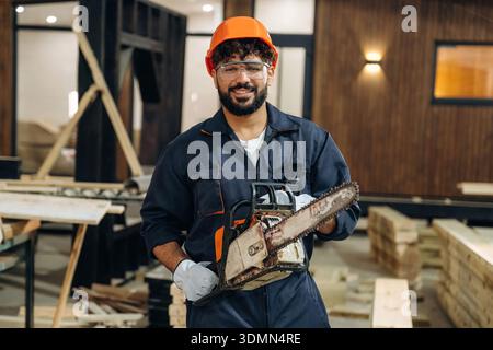 Powerful chainsaw, standing and holding it. Portrait of a male carpenter posing in a woodworking workshop. Stock Photo