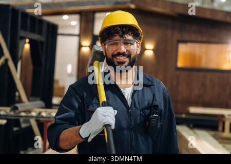 Big hammer is in hands. Portrait of a male carpenter posing in a woodworking workshop. Stock Photo