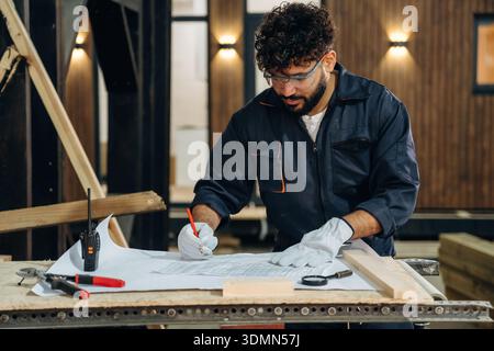 Plan is on the paper, using it. Male carpenter working with wooden materials in a professional workshop. Stock Photo