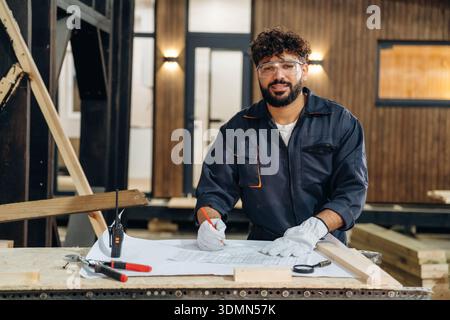 Plan is on the paper, using it. Male carpenter working with wooden materials in a professional workshop. Stock Photo