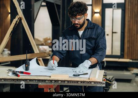 Plan is on the paper, using it. Male carpenter working with wooden materials in a professional workshop. Stock Photo