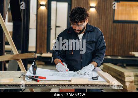 Plan is on the paper, using it. Male carpenter working with wooden materials in a professional workshop. Stock Photo