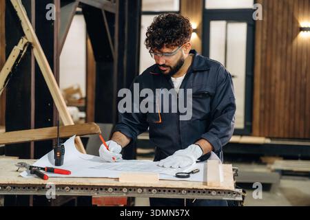 Plan is on the paper, using it. Male carpenter working with wooden materials in a professional workshop. Stock Photo
