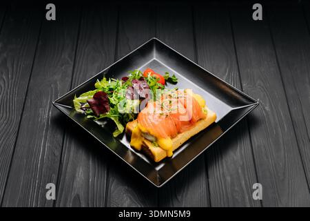 Salmon eggs benedict toast with hollandaise and green salad on black plate, dark table Stock Photo