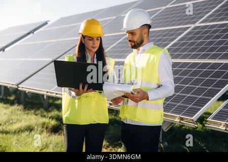 Laptop is in hands if woman. Two solar engineers standing together at a photovoltaic field, in bright safety vests. Stock Photo