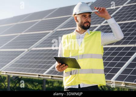 In yellow colored professional uniform. Male solar engineer examining photovoltaic panels at a power station. Stock Photo