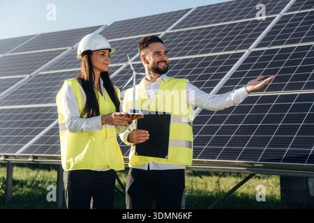 Windmill little model is in hands of woman. Two solar engineers standing together at a photovoltaic field, in bright safety vests. Stock Photo