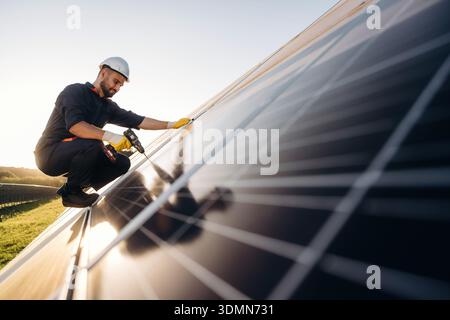 Service, sitting with drill and doing installation. Male solar engineer examining photovoltaic panels at a power station. Stock Photo