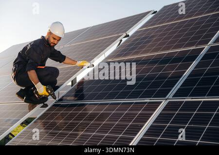Service, sitting with drill and doing installation. Male solar engineer examining photovoltaic panels at a power station. Stock Photo