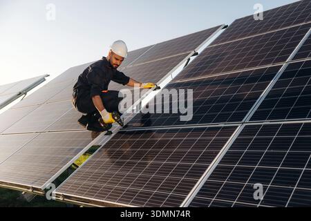 Service, sitting with drill and doing installation. Male solar engineer examining photovoltaic panels at a power station. Stock Photo