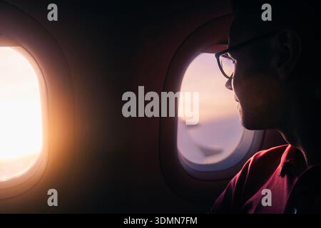 Passenger looking out airplane window at sunset during flight. View of glowing sky and horizon from aircraft. Solo travel, freedom, and emotional trav Stock Photo