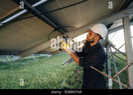 In white hardhat, using drill. Male solar engineer examining photovoltaic panels at a power station. Stock Photo