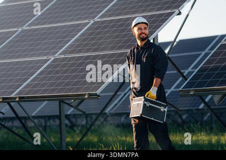 Ready for work, holding box with tools. Male solar engineer examining photovoltaic panels at a power station. Stock Photo