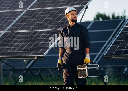 Ready for work, holding box with tools. Male solar engineer examining photovoltaic panels at a power station. Stock Photo