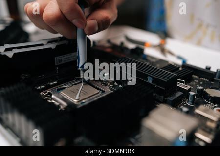Technician hand applying thermal paste to CPU of computer motherboard, demonstrating process of computer maintenance and repair in dark workshop. Stock Photo