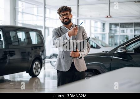Pen and notepad are in hands. Confident car salesman using digital tablet inside modern showroom. Stock Photo