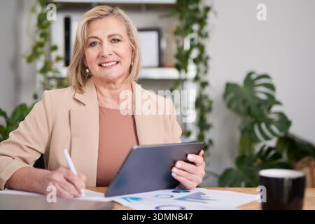 Professional Woman With Tablet Seated At Desk, Writing Documents In Modern Office Stock Photo