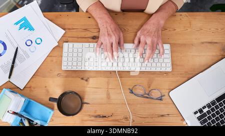 Business Professional Working At Wooden Desk With Keyboard, Laptop, Coffee, Documents, Graphs, And Glasses Stock Photo