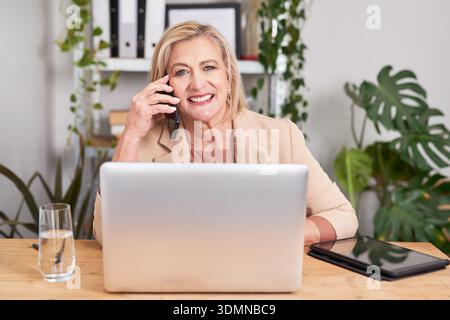 Businesswoman Smiles While On Phone At Desk With Laptop And Tablet In Modern Office Workspace Stock Photo