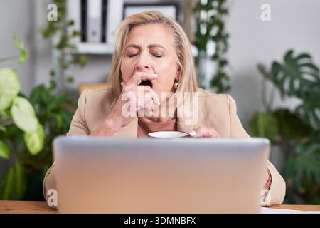 Business Woman Yawning At Laptop During Break With Coffee In A Green Office Stock Photo